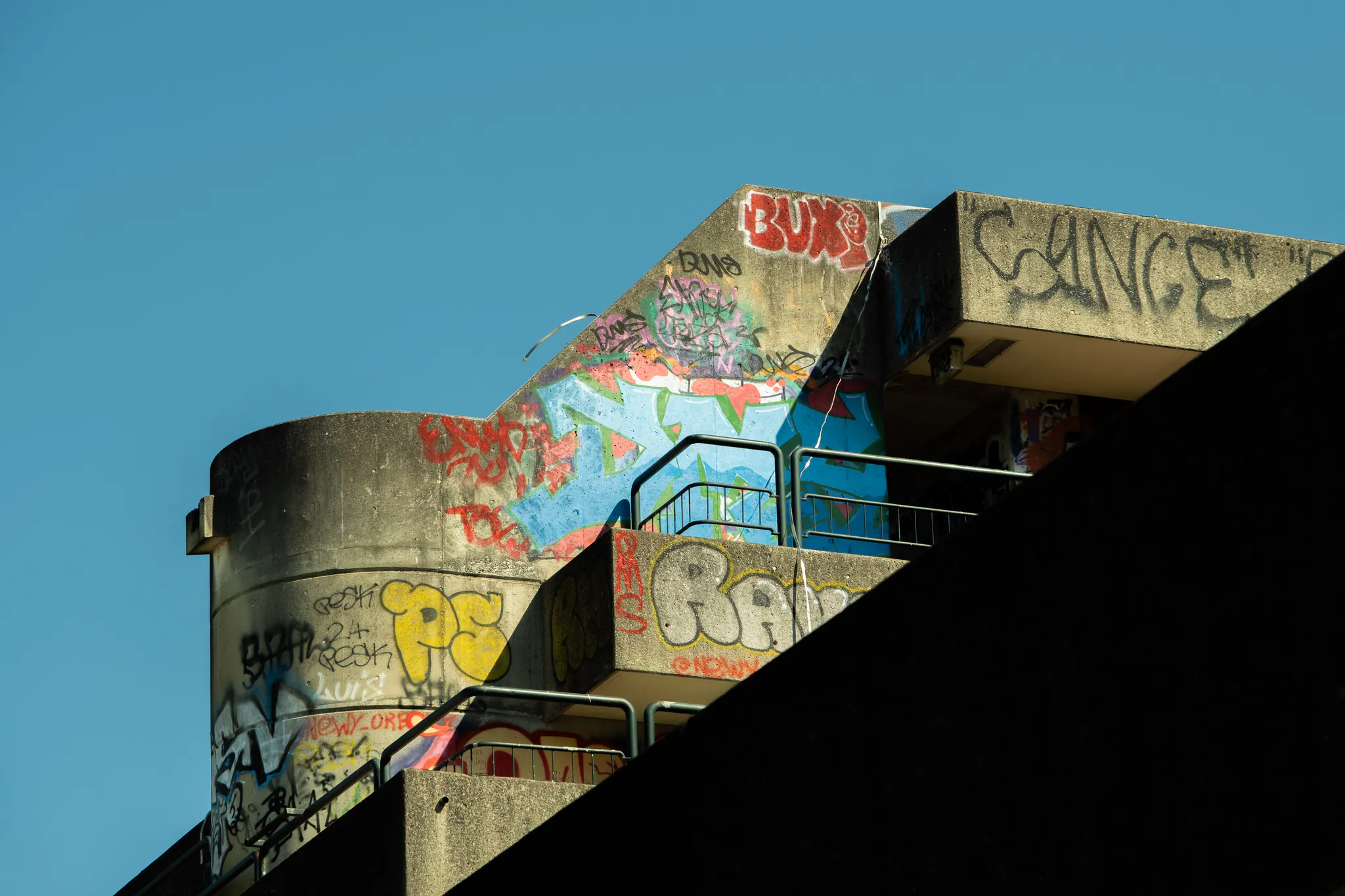the top of a concrete building against a clear blue sky, its curved walls and balconies layered in colourful graffiti