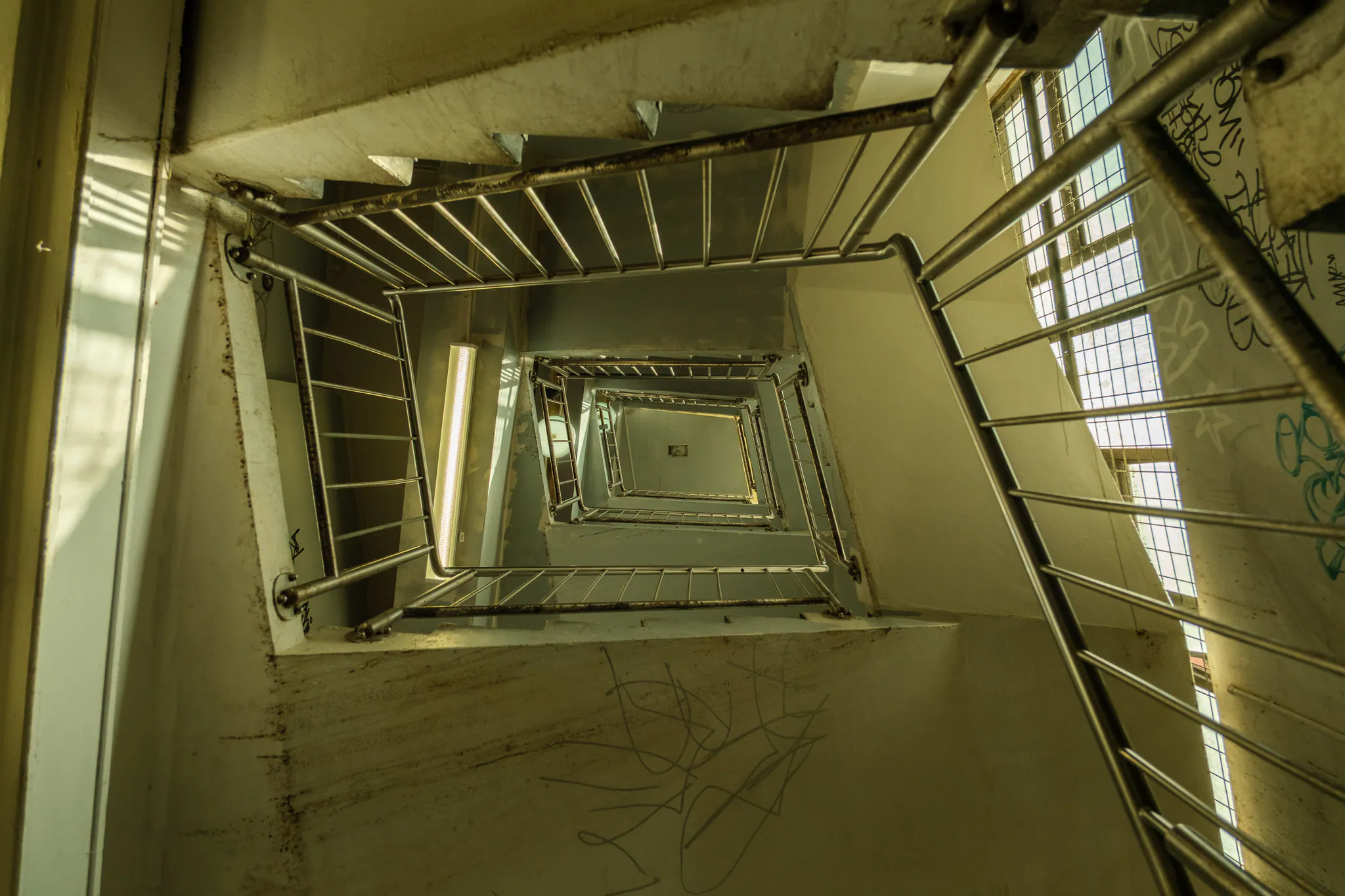 looking straight up a square stairwell, railings spiralling toward a skylight, graffiti on the walls