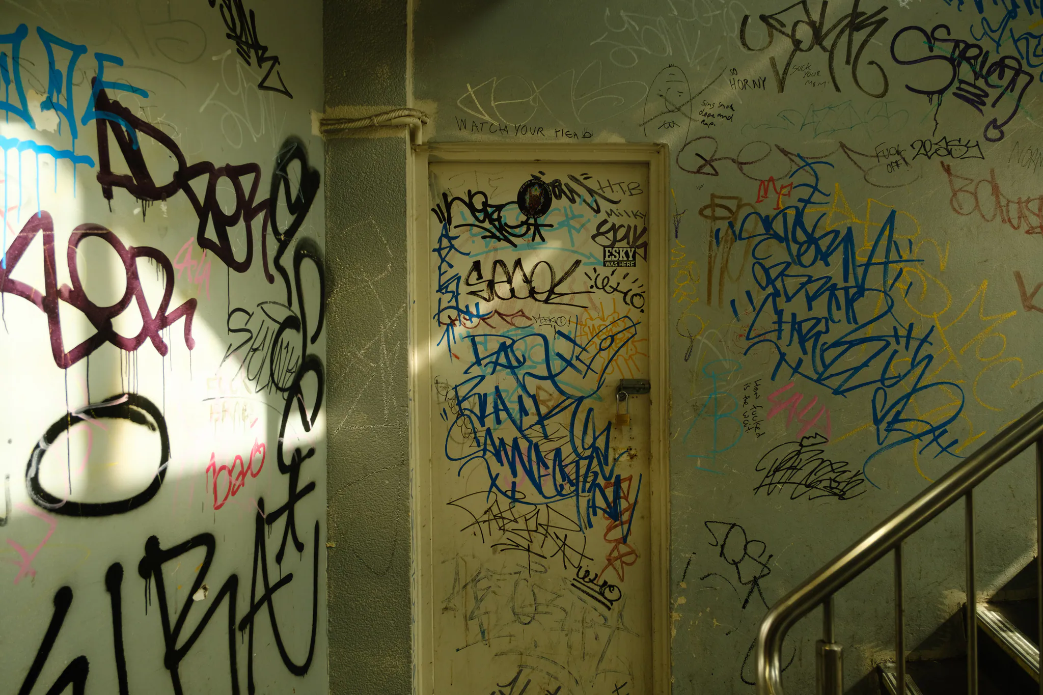 a door and walls completely covered in graffiti tags in a dim stairwell, a shaft of sun across the left wall
