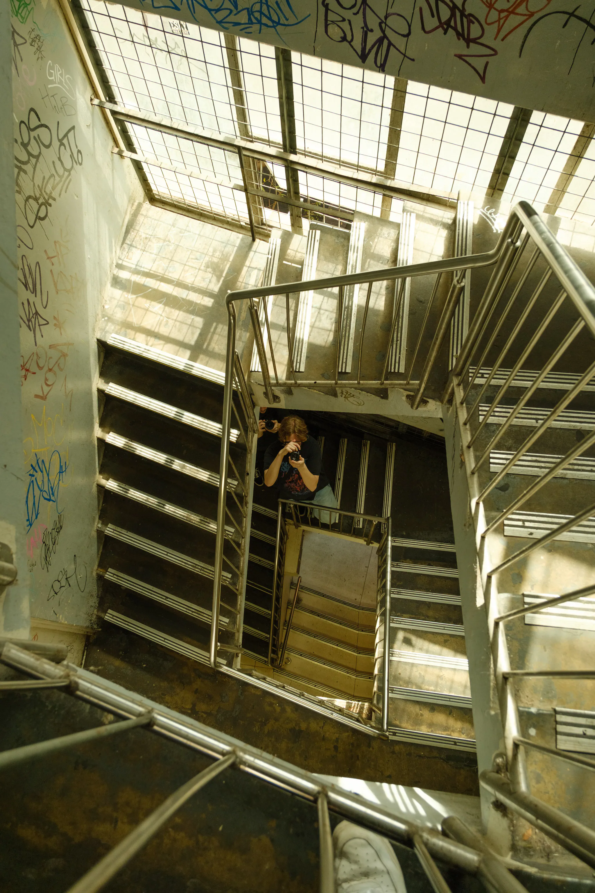 looking down a sunlit stairwell covered in tags, a friend with a camera framed at the bottom of the shaft