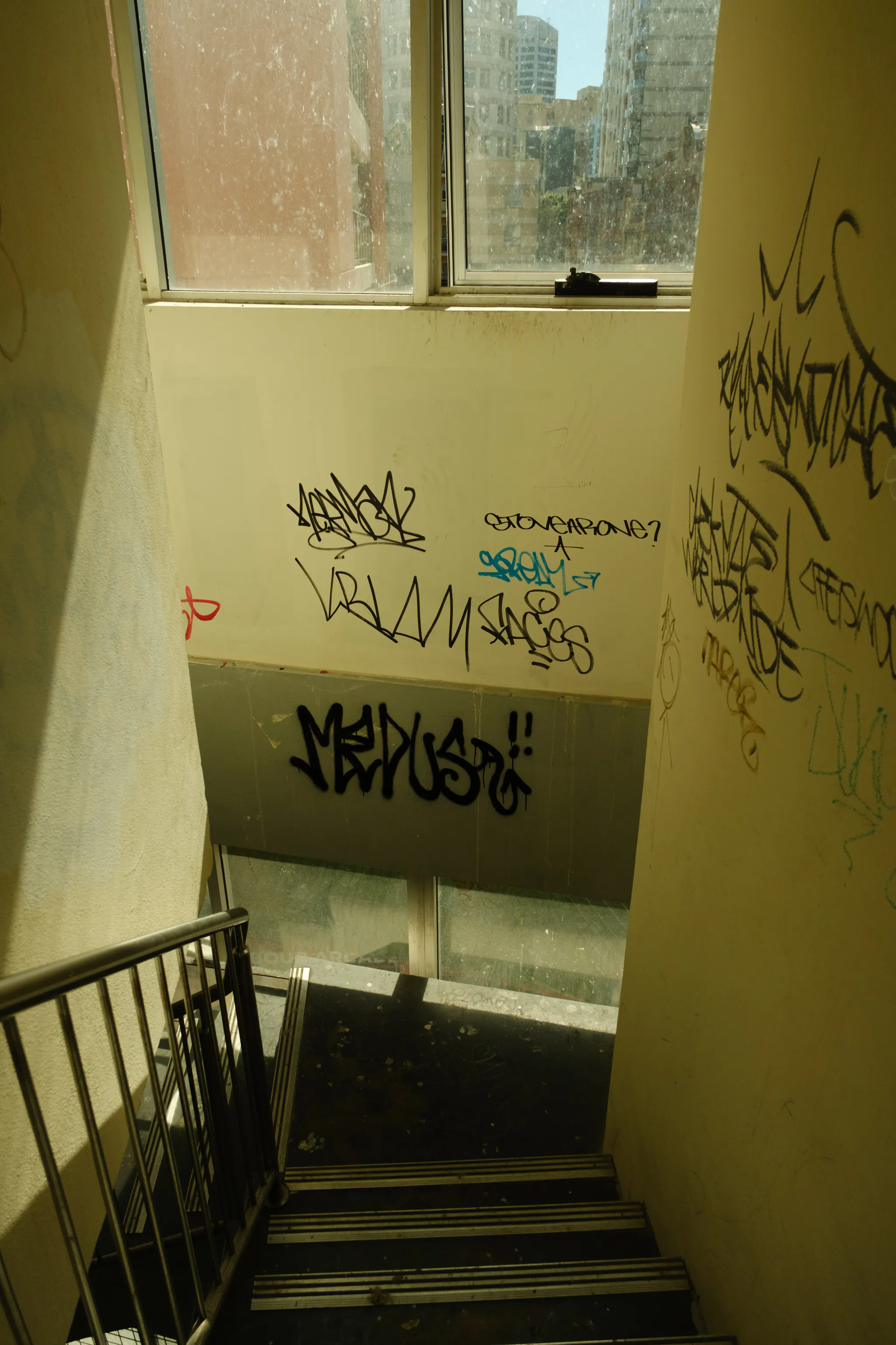stairs leading down to a landing with tagged walls and a dusty window looking out over city towers