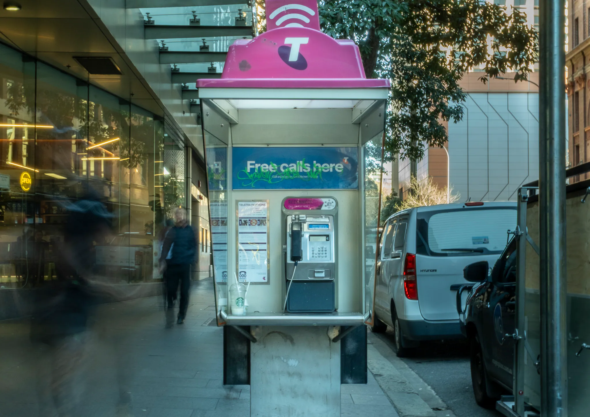 a telstra payphone with a magenta wifi roof outside a glass office lobby, one pedestrian blurred mid-stride, vans parked at the kerb