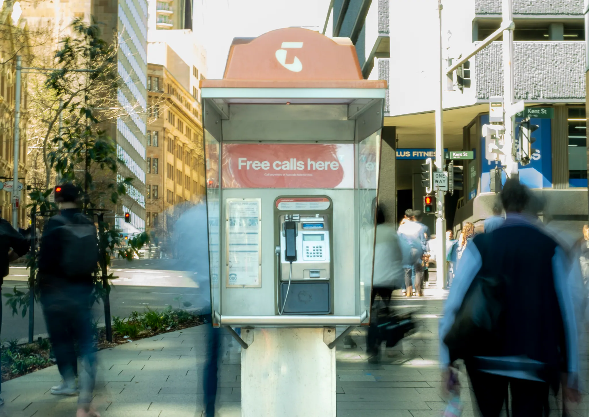 a pink telstra payphone centred on a sunny kent street corner while blurred pedestrians stream past on both sides