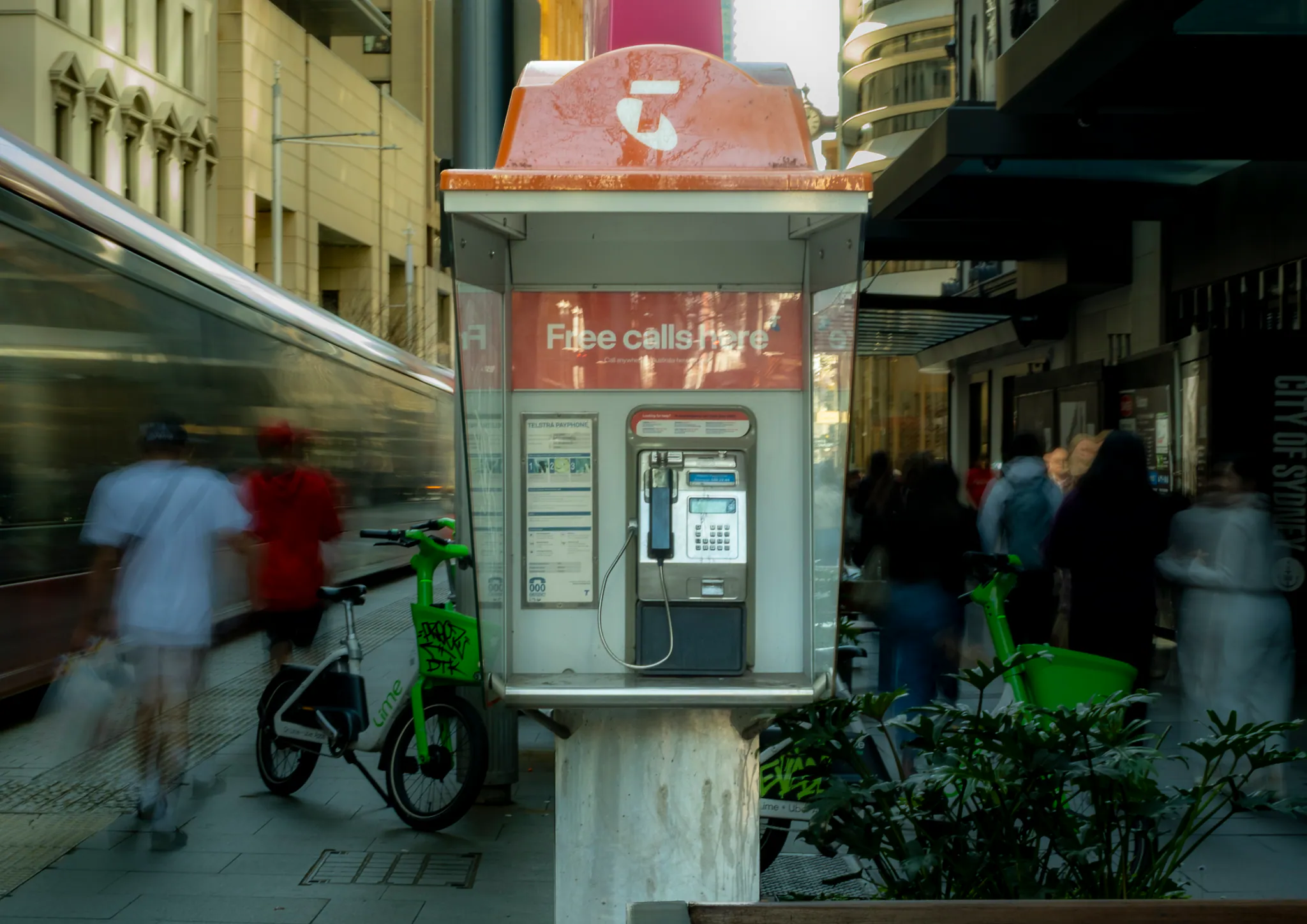 a payphone on a cbd footpath with a light rail tram smeared into a long blur behind it and lime e-bikes parked beside