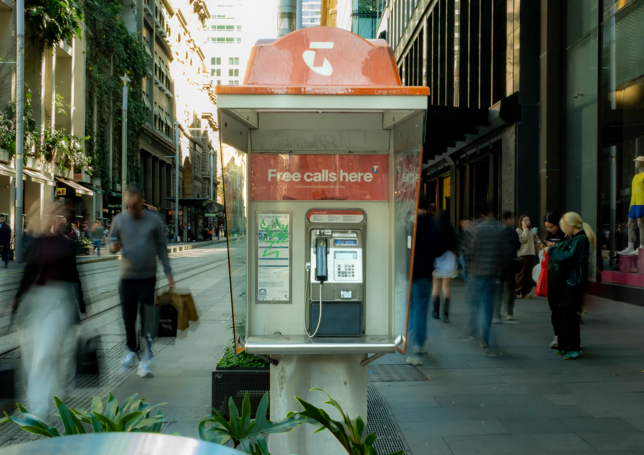 a payphone between planters on a busy pedestrian mall, shoppers blurred into ghosts as they walk around it