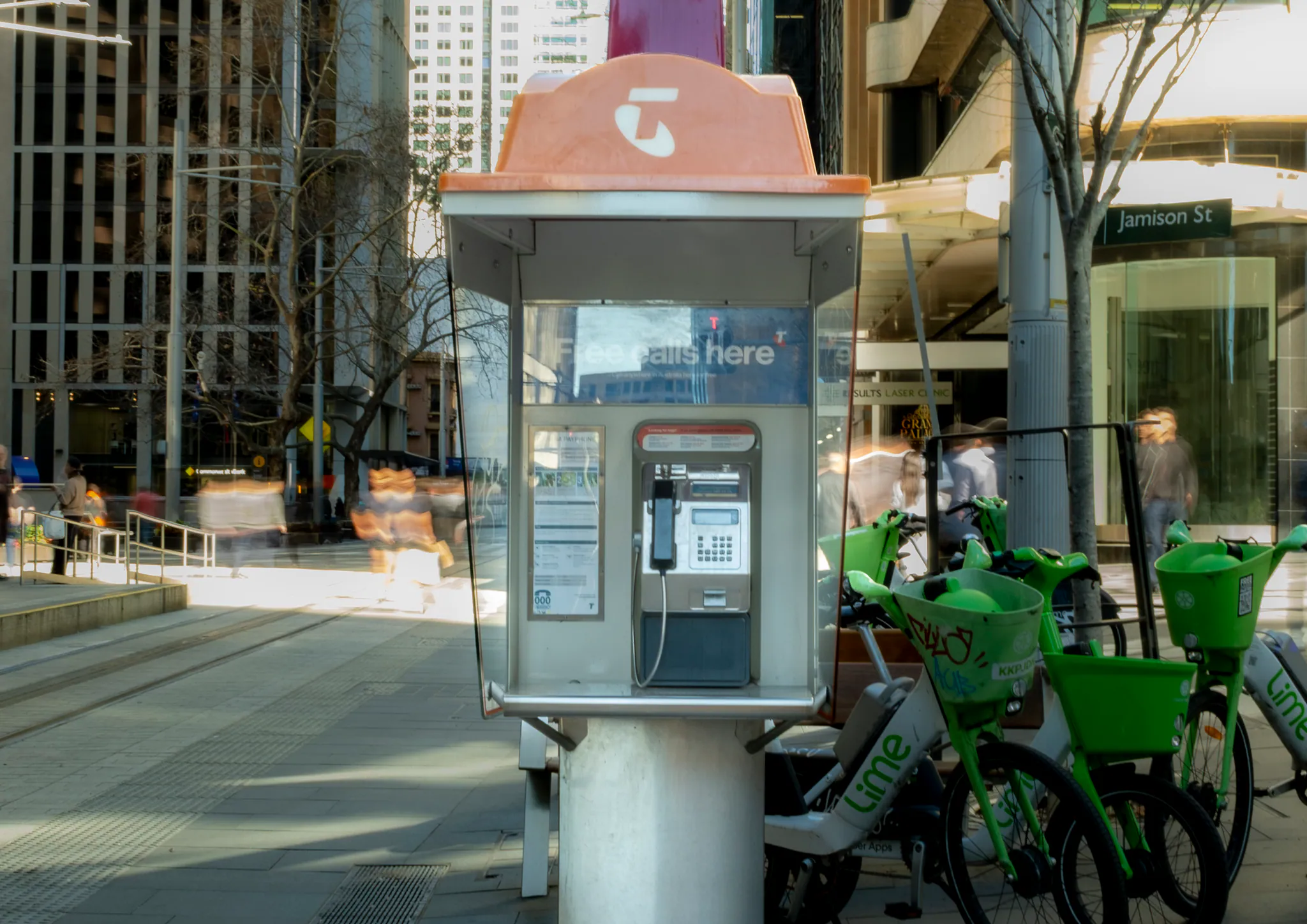 a payphone near jamison street beside a row of green lime e-bikes, sunlit commuters glowing into motion blur behind